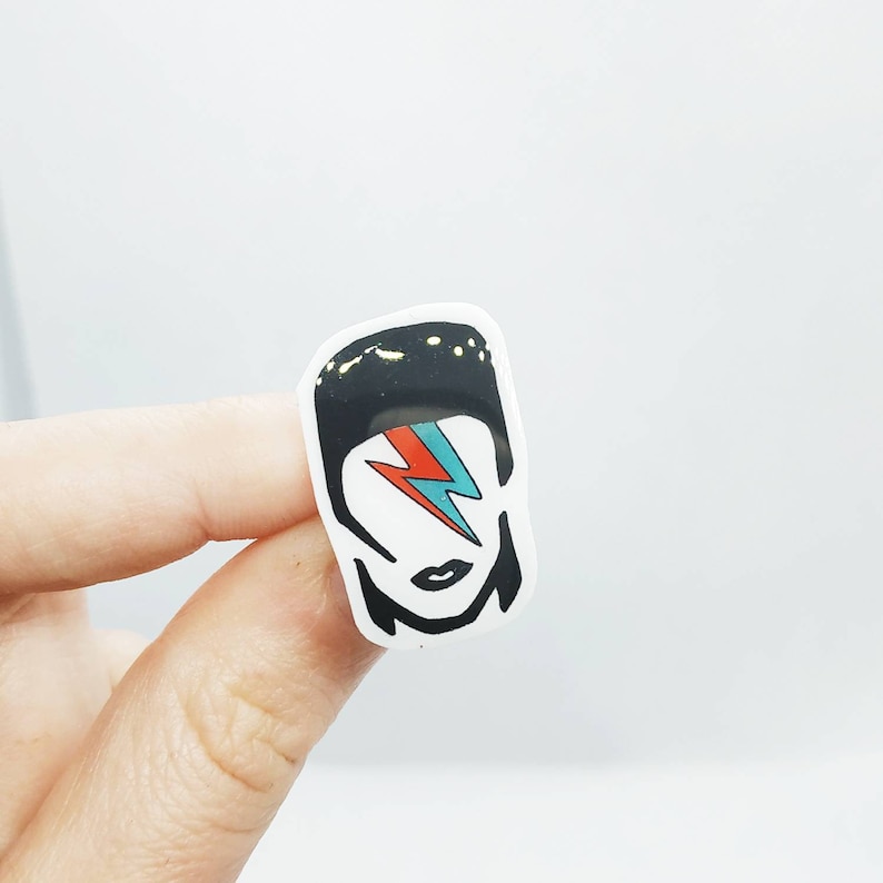 May include: A white sticker featuring a stylized illustration of a person with black hair and a red and blue lightning bolt across one eye. The sticker has a black outline and a white background.