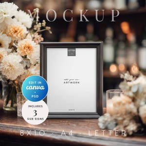 May include: A black framed mockup with the text "MOCKUP" at the top. The frame is on a wooden surface, with flowers on either side. The frame includes the text "add your own ARTWORK". The image also includes the text "EDIT IN canva + PSD" and "INCLUDES 3 SIZE SIGNS".