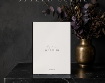 May include: A vertical white rectangular card mockup featuring the text add your own 5X7 DESIGN and DE-DI-TU. The card is positioned upright on a dark surface, with a brass vase containing dark dried foliage visible in the background.