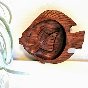 Puede incluir: Un cuenco de madera con forma de pez con un acabado marrón oscuro. El cuenco está tallado con detalles intrincados y tiene un borde ligeramente elevado.