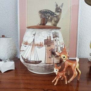 Puede incluir: Una jarra de galletas de cerámica con una tapa de madera marrón, que presenta una escena pintada de un velero y un castillo. Dos pequeñas figuritas de ciervos marrones están en primer plano.