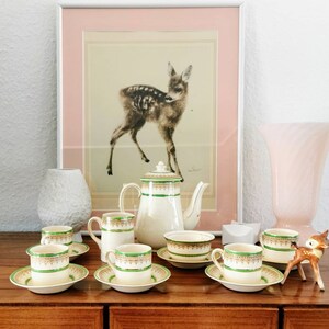 Könnte beinhalten: Ein Vintage-Teeservice mit grün-goldenen Verzierungen, darunter Tassen, Untertassen, eine Teekanne, ein Sahnekännchen und eine Zuckerdose. Ein gerahmter Hirschdruck und dekorative Gegenstände befinden sich im Hintergrund.