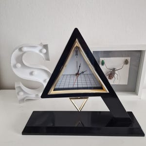 Puede incluir: Un reloj triangular negro con marco dorado y esfera blanca. El reloj tiene números romanos y un patrón de cuadrícula en la esfera. El reloj está sobre una base negra.