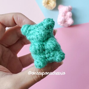 May include: A teal crocheted bear is held in a hand, with two more crocheted bears in yellow and pink in the background. The bears are on a pink and blue background. The text "@onceuponacheerio" is visible.