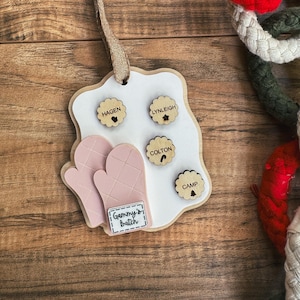 May include: A personalized wooden ornament with a white background, featuring four name tags and two pink oven mitts. The text "Gammy's Batch" is displayed on a small rectangular tag. The ornament is suspended by a burlap ribbon.