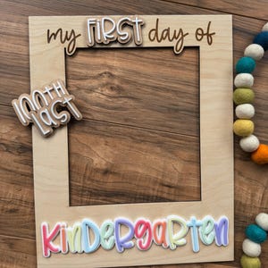 May include: A wooden photo frame with the words "my FIRST day of Kindergarten" in colorful 3D lettering. A smaller sign reads "100th Last". A colorful felt ball garland is in the background. The frame is on a wooden surface.