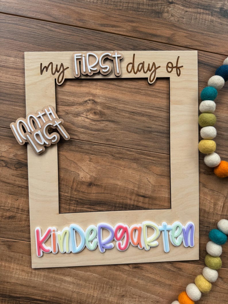 May include: A wooden photo frame with the words "my FIRST day of Kindergarten" in colorful 3D lettering. A smaller sign reads "100th Last". A colorful felt ball garland is in the background. The frame is on a wooden surface.