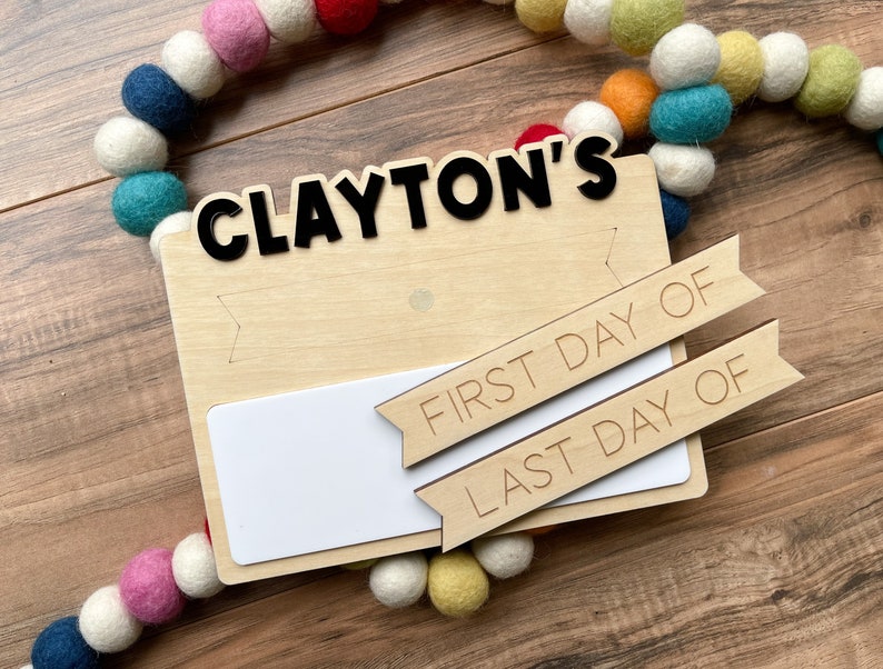 K&ouml;nnte beinhalten: Ein Holzschild mit dem Namen "CLAYTON'S" in schwarzen Buchstaben. Das Schild hat einen leeren Platz zum Schreiben und zwei Holzbanner, auf denen "ERSTER TAG VON" und "LETZTER TAG VON" steht.