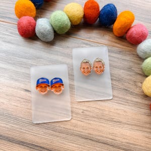 May include: Two pairs of personalized stud earrings featuring a smiling child's face. One pair shows the child wearing a blue baseball cap with the word "Blaze". Colorful felt balls in shades of orange, blue, yellow, and pink are arranged in a semi-circle.