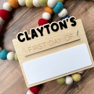 K&ouml;nnte beinhalten: Ein Holzschild mit dem Text "CLAYTON'S FIRST DAY OF" und einem leeren wei&szlig;en Feld zum Schreiben. Das Schild ist mit einer bunten Girlande aus Filzkugeln verziert.