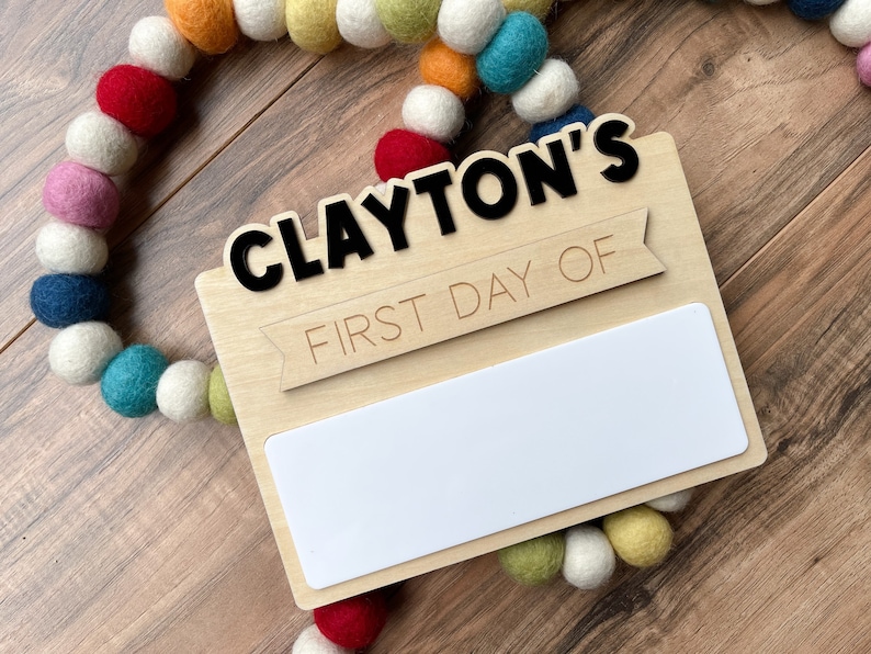 K&ouml;nnte beinhalten: Ein Holzschild mit dem Text "CLAYTON'S FIRST DAY OF" und einem leeren wei&szlig;en Feld zum Schreiben. Das Schild ist mit einer bunten Girlande aus Filzkugeln verziert.
