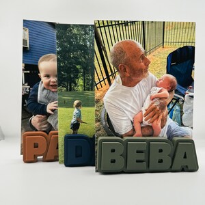 May include: Three vertical photo displays featuring custom block letter bases. The left base is orange with the letters PAD, the center is blue with the letter D, and the right base is olive green with the letters BEBA, each holding a personal photograph.
