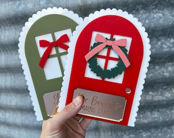 May include: Two decorative fairy doors, one red and one green, with white scalloped edges. The red door has a pink bow and green wreath, while the green door has a red bow. Both doors have a gold nameplate that reads "The Bourne's MAGIC DOOR".