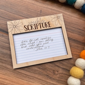 May include: Wooden frame with the words "weekly SCRIPTURE" and a floral design. Inside is a lined index card with a handwritten Bible verse from John 13:7. A felt ball garland in various colors is in the background.