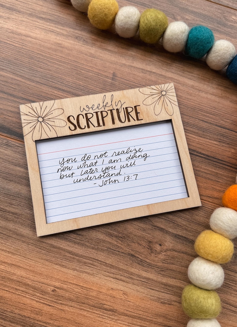 Może przedstawiać: Drewniana ramka z napisem "weekly SCRIPTURE" i kwiatowym wzorem. Wewnątrz znajduje się liniowana karta indeksu z ręcznie napisanym wersetem biblijnym z Jana 13:7. Girlanda z filcowych kulek w r&oacute;żnych kolorach znajduje się w tle.
