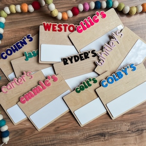 K&ouml;nnte beinhalten: Ein Satz von acht Holz-Namensschildern mit bunten Buchstaben, die Namen wie "Cohen's", "Ryder's" und "Colby's" buchstabieren. Jedes Schild hat einen wei&szlig;en Bereich zum Schreiben oder Zeichnen.