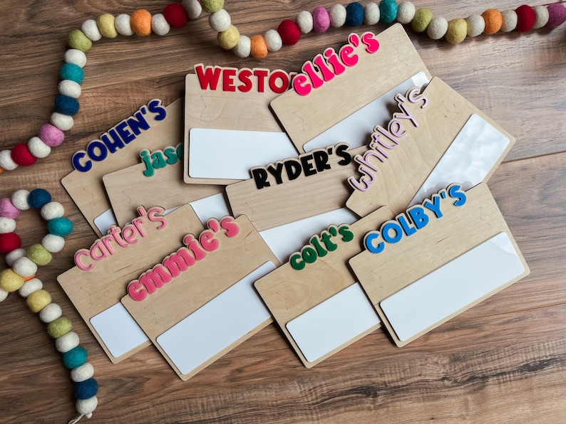 K&ouml;nnte beinhalten: Ein Satz von acht Holz-Namensschildern mit bunten Buchstaben, die Namen wie "Cohen's", "Ryder's" und "Colby's" buchstabieren. Jedes Schild hat einen wei&szlig;en Bereich zum Schreiben oder Zeichnen.