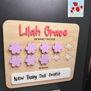 May include: Wooden reward tracker with the name "Lilah Grace" in pink 3D letters. The tracker has pink flower-shaped tokens and the text "New Baby Doll Outfit" on a white label. A small painting of red flowers is visible.