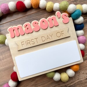 K&ouml;nnte beinhalten: Ein Holzschild mit dem Text "mason's first day of" in rosa Buchstaben. Das Schild hat einen leeren wei&szlig;en Bereich zum Schreiben des Datums.