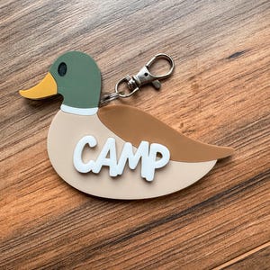 May include: A duck-shaped keychain with a green head, yellow beak, and brown wing. The word "CAMP" is in white letters on the duck's body. It has a silver-colored clasp for attaching to keys or a bag. The background is a wooden surface.
