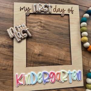 May include: A wooden photo frame with the words "my FIRST day of" and "Kindergarten" in colorful, layered lettering. The frame also includes a smaller sign that says "100th LAST". A colorful felt ball garland is visible in the background.