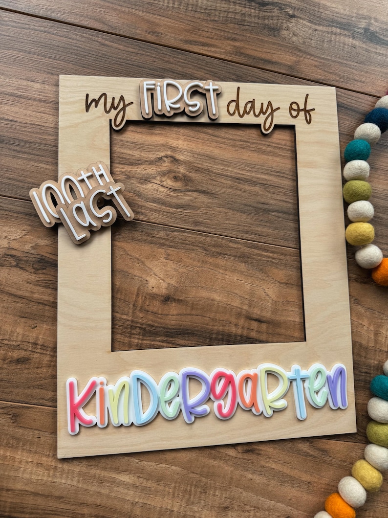 May include: A wooden photo frame with the words "my FIRST day of" and "Kindergarten" in colorful, layered lettering. The frame also includes a smaller sign that says "100th LAST". A colorful felt ball garland is visible in the background.