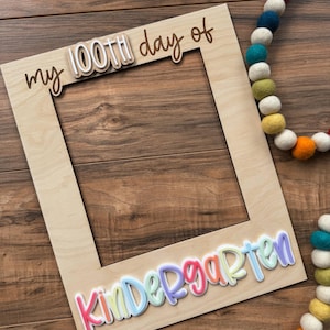 May include: A wooden photo frame with the words "my 100th day of" and "Kindergarten" in colorful, layered lettering. A colorful felt ball garland is in the background. The frame is made of light-colored wood.