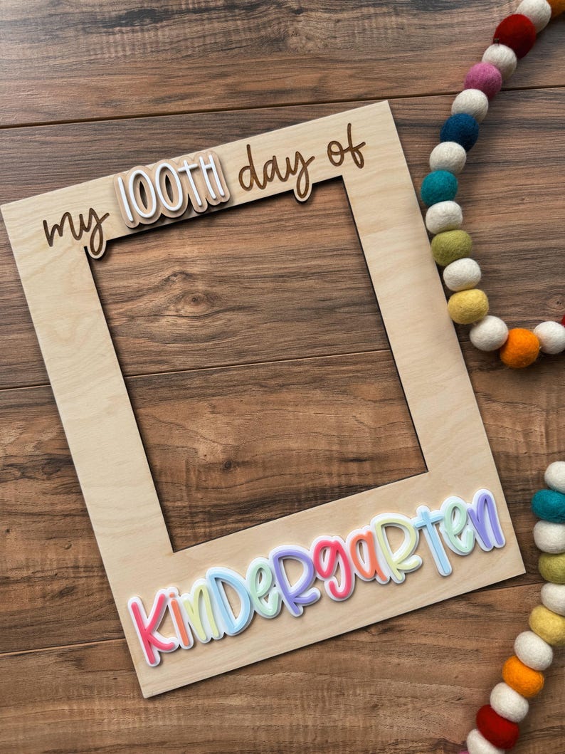 May include: A wooden photo frame with the words "my 100th day of" and "Kindergarten" in colorful, layered lettering. A colorful felt ball garland is in the background. The frame is made of light-colored wood.