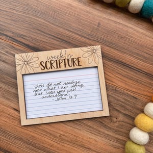 Może przedstawiać: Drewniana ramka na cotygodniowe Pismo Święte z odręczną notatką. Ramka ma kwiatowe akcenty i napis "weekly SCRIPTURE". Notatka brzmi: "You do not realize now what I am doing but later you will understand. - John 13:7".