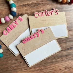 K&ouml;nnte beinhalten: Drei Holzschildchen mit wei&szlig;en Fl&auml;chen zum Beschreiben. Jedes Schildchen tr&auml;gt einen rosafarbenen Namen: Carly's, Emery's und Carter's.