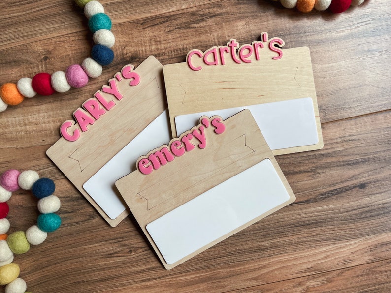 K&ouml;nnte beinhalten: Drei Holzschildchen mit wei&szlig;en Fl&auml;chen zum Beschreiben. Jedes Schildchen tr&auml;gt einen rosafarbenen Namen: Carly's, Emery's und Carter's.
