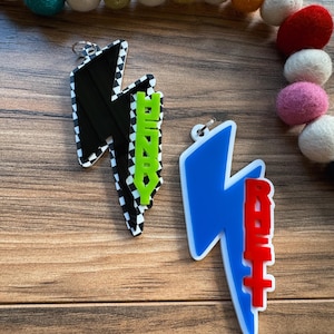 May include: Two lightning bolt-shaped keychains. One is black with a checkered pattern and the word "HENRY" in green. The other is blue with the word "RENT" in red. Both have silver key rings.