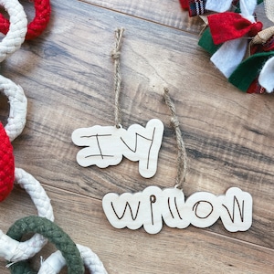 Może przedstawiać: Dwa drewniane ozdoby z napisami "Ivy" i "Willow" napisanymi czarnym markerem. Ozdoby wiszą na sznurku i znajdują się na drewnianym tle.