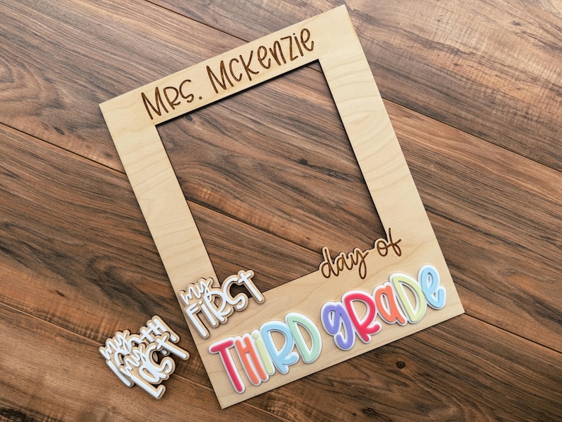 May include: A wooden photo frame with the words "Mrs. McKenzie" engraved at the top. The phrase "My First Day of Third Grade" is written in colourful, 3D lettering. The frame is made of light-coloured wood.