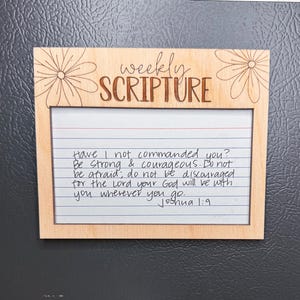 Może przedstawiać: Drewniana ramka z napisem "weekly SCRIPTURE" i ręcznie napisanym wersetem biblijnym z Księgi Jozuego 1:9. Ramka ma kwiatowy wz&oacute;r i jest wyświetlana na szarej powierzchni.