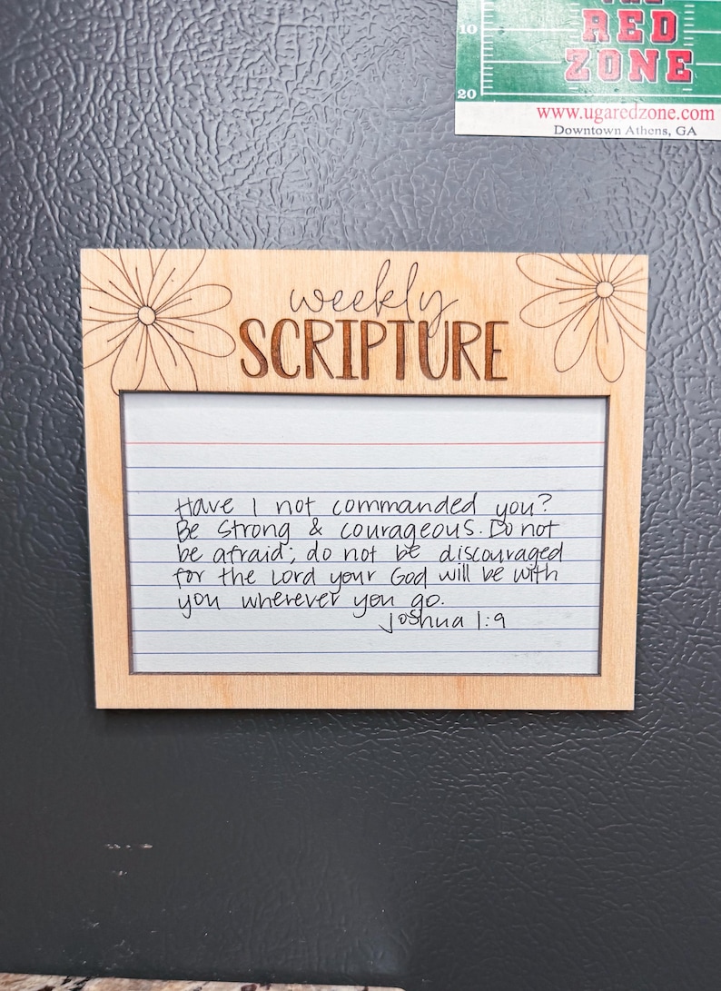 Może przedstawiać: Drewniana ramka z napisem "weekly SCRIPTURE" i ręcznie napisanym wersetem biblijnym z Księgi Jozuego 1:9. Ramka ma kwiatowy wz&oacute;r i jest wyświetlana na szarej powierzchni.