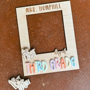 May include: A wooden photo frame with the text "MRS. HEMPHILL" at the top. Below, the words "MY FIRST" and "day of THIRD GRADE" are attached. A separate piece reads "100th day". The frame has a natural wood finish.