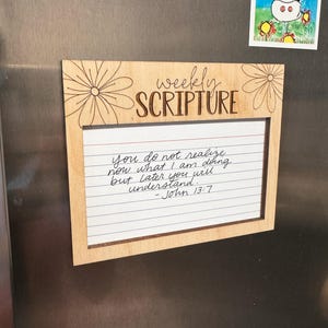Może przedstawiać: Drewniana tablica z cotygodniowym pismem z napisem "weekly SCRIPTURE". Tablica ma białą, liniowaną wkładkę papierową z odręcznym tekstem z Jana 13:7. Ozdobne elementy kwiatowe są wytrawione w drewnie. Powyżej tablicy przymocowany jest magnes z rysunkiem krowy.