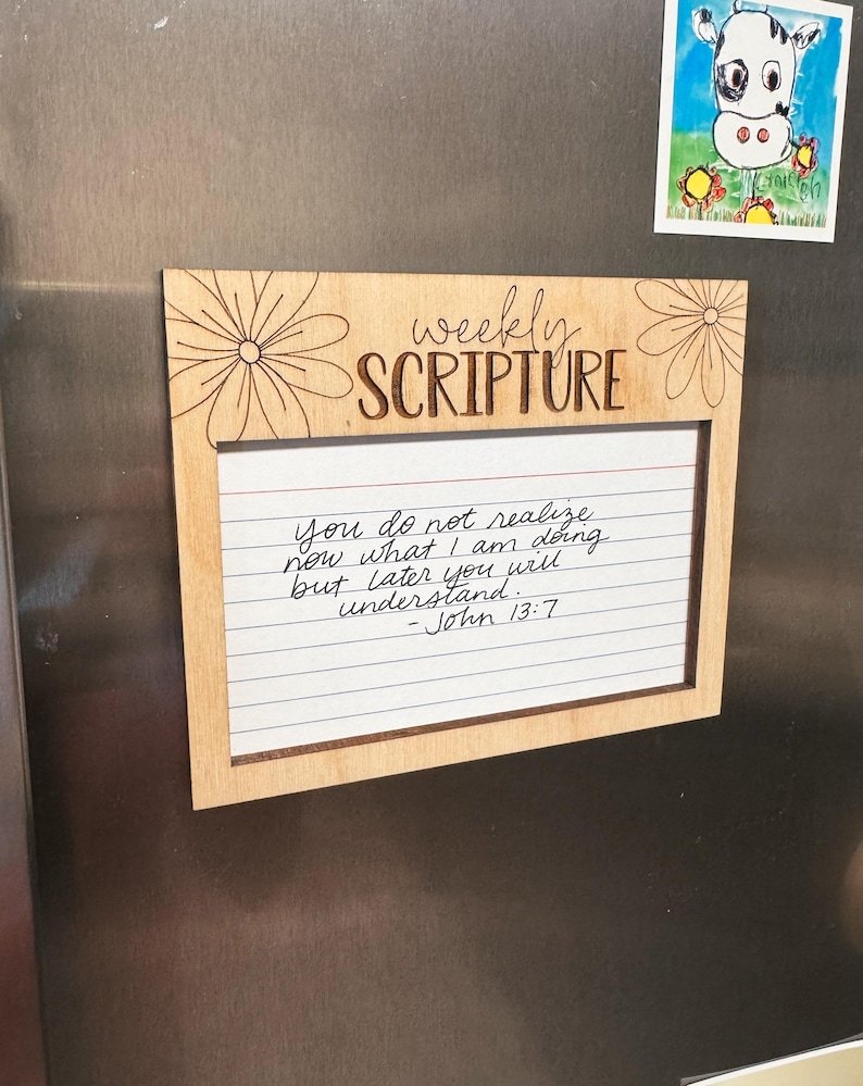 Może przedstawiać: Drewniana tablica z cotygodniowym pismem z napisem "weekly SCRIPTURE". Tablica ma białą, liniowaną wkładkę papierową z odręcznym tekstem z Jana 13:7. Ozdobne elementy kwiatowe są wytrawione w drewnie. Powyżej tablicy przymocowany jest magnes z rysunkiem krowy.