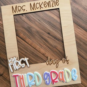 May include: A light-coloured wooden photo frame with the words "Mrs. McKenzie" engraved at the top. The phrase "my first day of THIRD GRADE" is written across the bottom in colourful, raised letters.