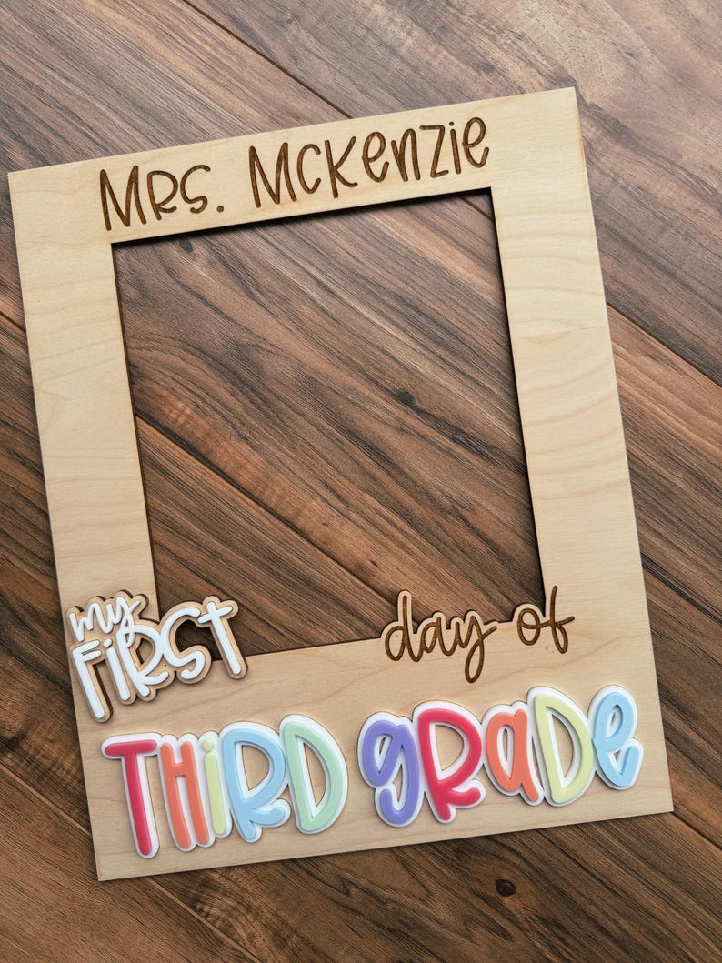May include: A light-coloured wooden photo frame with the words "Mrs. McKenzie" engraved at the top. The phrase "my first day of THIRD GRADE" is written across the bottom in colourful, raised letters.
