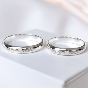 Puede incluir: Dos anillos de boda plateados con bordes redondeados. Cada anillo tiene los nombres grabados "mos" y "matt" separados por un pequeño corazón. Los anillos se muestran sobre una superficie blanca.