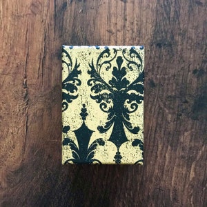 May include: A small, square box with a black and yellow damask pattern. The box is made of cardboard and has a matte finish.