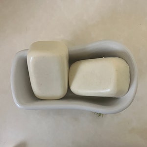 May include: Two white bars of soap in a white ceramic soap dish shaped like a bathtub.