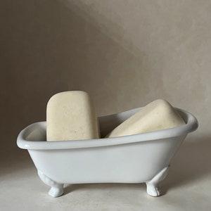 May include: Two white bars of soap in a white porcelain bathtub-shaped soap dish with three feet.