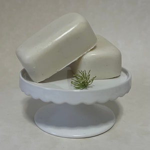 May include: Two white bars of soap on a white pedestal cake stand with a small green plant on top.
