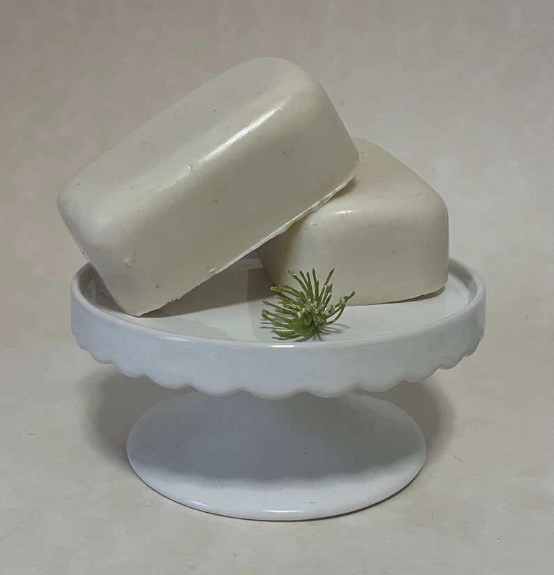 May include: Two white bars of soap on a white pedestal cake stand with a small green plant on top.