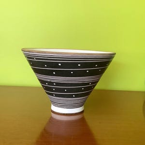 May include: A black and white ceramic bowl with a distinctive shape. The bowl has horizontal white stripes and white polka dots. The rim has a brown band. The bowl is on a wooden surface.