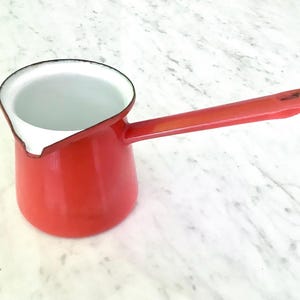 Puede incluir: Una cafetera de esmalte rojo brillante con un mango largo y un interior blanco. La cafetera presenta un pequeño pico vertedor en el borde y un ribete negro a lo largo del borde superior. La superficie exterior es lisa, brillante y roja.