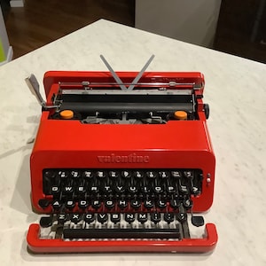 Könnte beinhalten: Eine leuchtend rote Valentine-Schreibmaschine mit schwarzen Tasten und dem Wort "valentine" auf der Vorderseite. Die Schreibmaschine hat einen silbernen Hebel und eine Papierführung. Die Tasten sind in einem QWERTZ-Layout angeordnet.